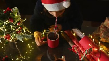 Noel Baba şapkalı bir çocuk Noel ışıkları, yeşillik ve paketlenmiş hediyeler arasında bardaktan su içer..