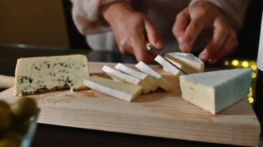 Kadın tahta kesme tahtasıyla peynir dilimliyor. Yumuşak ışıklandırmayla peynir kesen ellerin yakın çekimi. Arka planda zeytin ve mavi peynir görünür..