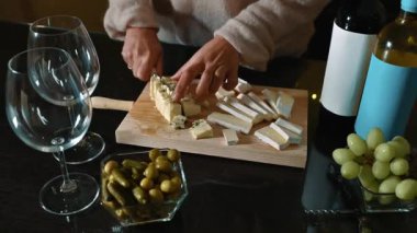 Tahtada peynir kesen bir kadın. İki şarap kadehi, zeytin, üzüm ve iki şarap şişesi var. Romantik bir akşam yemeği hazırlıyorum..