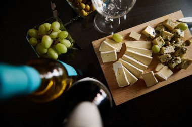 Zarif bir peynir tabağında üzüm ve şarapla birlikte çeşitli peynirlerden dilimler yer alıyor. Tahtaya yerleştirilmiş bu düzenek, toplanma ve şarap tatma deneyimleri için mükemmel..