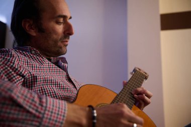 A close-up of a man playing an acoustic guitar indoors, conveying focus and passion. The ambient lighting creates a warm and relaxed atmosphere celebrating the art of music. clipart