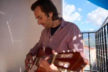 A man playing an acoustic guitar on a balcony with a scenic outdoor background. The day is clear with blue skies, creating a serene and relaxed atmosphere perfect for music and contemplation. clipart