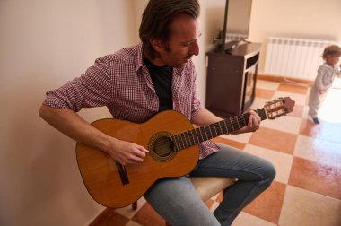 A man seated in a living room plays an acoustic guitar while a child is nearby. The scene captures a warm family moment with music and relaxation in a home environment. clipart