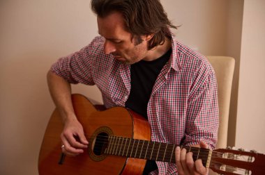 Huzurlu bir ortamda oturan bir adam konsantrasyon ve hassasiyetle akustik gitar çalar..