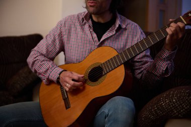 A man wearing a plaid shirt plays an acoustic guitar in a living room setting, showcasing a casual and intimate atmosphere. clipart