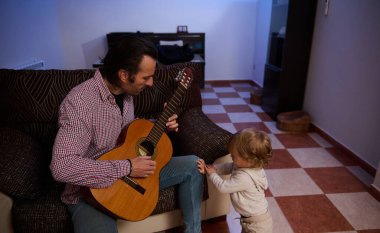 A man is playing guitar in a cozy living room while a small child attentively watches him, showcasing a wholesome family moment filled with music, bonding, and a shared interest. clipart