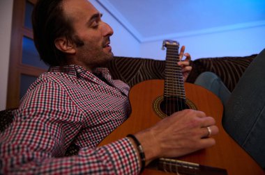 A man casually strumming an acoustic guitar indoors, wearing a plaid shirt, enjoying music. The cozy setting and warm lighting emphasize relaxation and a passion for musical performance. clipart