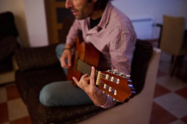 Kanepede oturmuş, evde akustik gitar çalan birinin yakın çekimi. Müzik, rahatlama, yaratıcılık ve rahat bir ortamda hobilerin tadını çıkarmak için mükemmel bir yöntem..