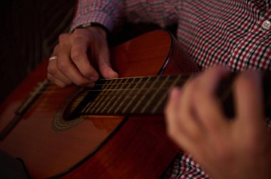 Kapalı mekanda ahşap akustik gitar çalan birinin ellerinin yakın çekimi. Sahne, gitarın tellerine ve detaylarına odaklanarak müzik yaratmanın sıcaklığını ve sanatsallığını yakalıyor..