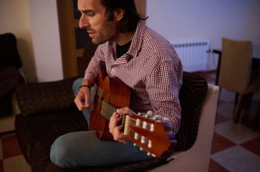 Bir adam evde akustik gitar çalıyor, günlük kıyafetler giyiyor, sıcak bir atmosfer yaratıyor..