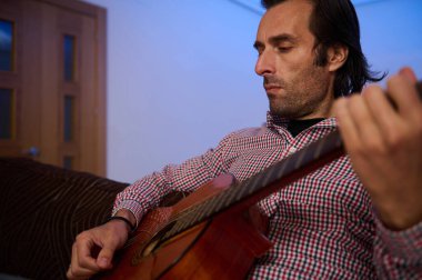 Kapalı bir koltukta oturan bir adam, ahşap akustik gitar çalarken derinden odaklanmış..