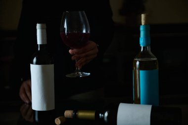 Şarap şişeleri ve kırmızı şarap kadehini tutan bir elin yer aldığı düzenleme zarif ve kutlama ortamını yansıtıyor..