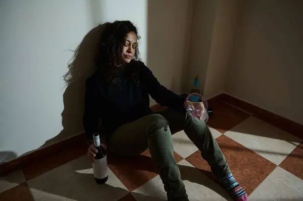 Sıkıntılı bir yüzü olan bir kadın şarap şişesi ve cam tutarak duvara yaslanır. Ayarlar yalnızlık, tefekkür ya da kişisel zorluklarla mücadele içeriyor..