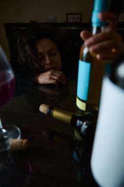Bir kadın üzüntüsünü ifade eder, masada bir şarap şişesine uzanır, alkol bağımlılığı ve onun duygusal bilançosuyla ilgili mücadeleleri vurgular..