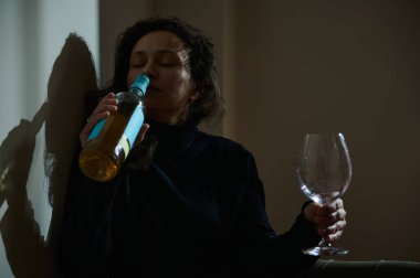 Bir kadın şarap şişesi ve bardakla yere oturmuş yalnızlık, içtenlik ve alkol tüketimiyle mücadele konularını vurguluyor..