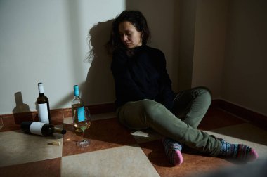 A woman sits on a tiled floor with wine bottles and a glass, suggesting complexities. clipart