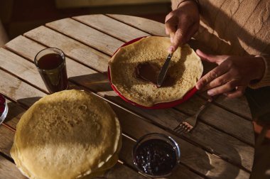 Bir insan reçel ve içeceklerle birlikte çikolata yayarak krep hazırlar. Doğal ışık altında ahşap bir masada yakalanan bu resim, sıcak, lezzetli ve rahatlatıcı bir yemeğin özünü yakalar..