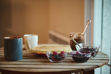 Krep, böğürtlen, reçel kavanozları ve ahşap bir masada sıcak içeceklerin yer aldığı samimi bir kahvaltı sahnesi davetkar ve kırsal bir sabah yemeği konseptini yakalıyor..