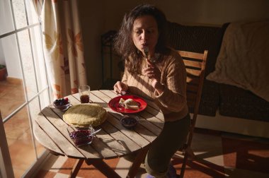 Bir kadın ahşap bir masada oturmuş, içinde krep ve reçel bulunan bir tabak kahvaltının tadını çıkarıyor. Güneş ışığı pencereden geçer ve rahat bir yemek ortamı yaratır..