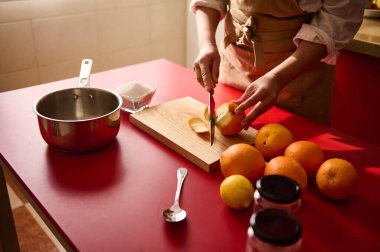 A person skillfully slices citrus fruits on a wooden board, ready for culinary preparation. The vibrant setup includes oranges, a saucepan, and jars, showcasing a clean and inviting cooking atmosphere. clipart