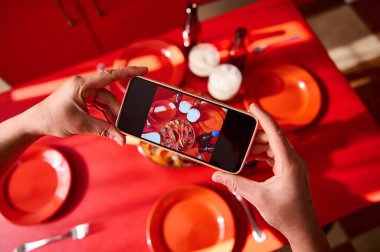 Akıllı telefonu tutan eller, parlak bir masanın üstünde yemeğin tepeden tırnağa fotoğrafını çekiyor. Resim, kırmızı temalı arka planda modern fotoğrafçılık, aşçılık sanatı ve canlı estetiği vurguluyor..