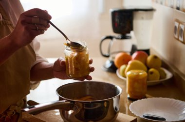 A close-up of a jar being filled with homemade preserves in a cozy kitchen setting. Various fresh fruits and kitchen utensils are seen in the background, creating a warm and inviting atmosphere. clipart