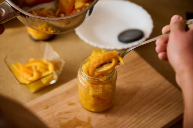 Bir el, kavanozu yeni pişmiş portakal kabuklarıyla doldurmak için çatal kullanır. Mutfak ortamında ahşap bir kesme tahtasındaki mutfak reçellerini hazırlar..