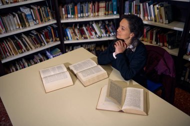 Bir kadın kütüphane masasında oturur, etrafı açık kitaplarla çevriliyken dikkatle başka tarafa bakar. Sahne, entellektüel merak, yansıma ve bir kütüphanede öğrenme ve çalışma atmosferini taşır.