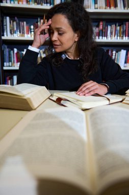 Bir öğrenci kütüphanede dikkatlice çalışır, açık kitaplarla çevrilidir ve notlar alır. Bu resim, akademik bir ortamda odak, eğitim ve araştırma atmosferini yakalar..