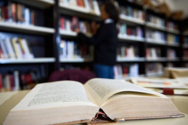 An open book lies on a table in a library, with bookshelves and a reader in the background.