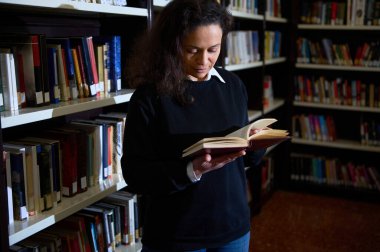 A person fully engrossed in reading a book while standing in a well-organized library, surrounded by bookshelves filled with a variety of colorful books. clipart