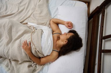 Yatakta huzur içinde yatan genç bir kadın, yukarıdan yakalanmış, dokulu bir battaniyeye sarılmış..