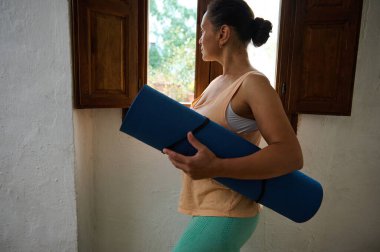 A woman stands in a minimalist room with wooden windows, holding a yoga mat while gazing outside. The scene depicts a calm and introspective moment, emphasizing a lifestyle focused on fitness and mindfulness. clipart