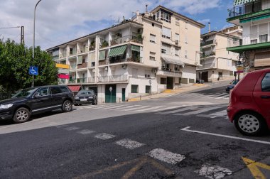 Park halindeki araçların ve konut binalarının bulunduğu eğimli arazili bir cadde. Yerleşim, kenar mahalle sakinliği ve doğal unsurların karışımı ile taşınıyor.