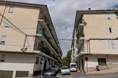 İki konut binası arasında yer alan bir cadde. Balkonları, park edilmiş arabaları ve görünür elektrik kabloları, parçalı bulutlu bir gökyüzünün altında..