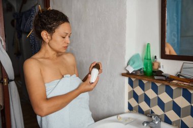 Havluya sarılmış bir kadın banyo lavabosunda kozmetik şişesini inceliyor..