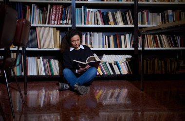 A young woman enjoying reading a book while sitting by a bookshelf in a library. The atmosphere is calm and intellectual, portraying the joy of literature and the serenity of learning.