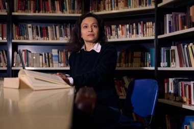 Bir kadın kütüphanede bir masada oturuyor, dikkatle büyük bir kitap okuyor. Arkaplanda çeşitli ciltlerle dolu kitaplıklar yer alıyor, öğrenme ve içsel bir ortam yaratıyor.