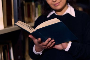 A person holding and reading a book while standing near shelves in a library, showcasing a quiet and studious atmosphere, perfect for learning, research, or personal development. clipart