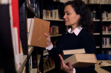 A woman holding several books while selecting another book from the shelf in a library, expressing a love for reading and education in an organized and peaceful environment.