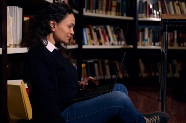 Kütüphanenin zemininde oturan bir kadın, kitaplıklarla çevrili bir dizüstü bilgisayarla çalışıyor. Ayar, öğrenmeyi, sessiz konsantrasyonu ve bilgi keşiflerinde teknolojinin kullanımını vurguluyor..