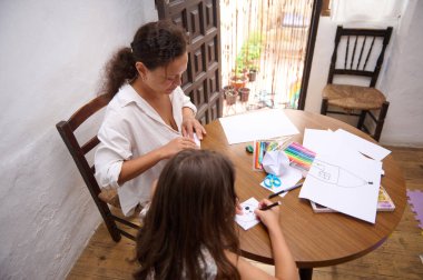 Bir anne ve kız evde bir sanat etkinliğine katılıyor, yaratıcılığı teşvik ediyor ve renkli malzemelerle ahşap bir masa üzerinde çizim ve el sanatları ile bağ kuruyor..