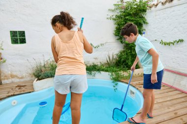 A woman and a child cleaning a backyard pool with nets on a wooden patio. clipart