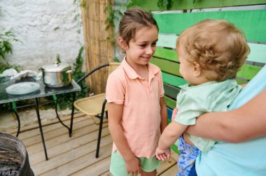Genç bir kız ve yeni yürümeye başlayan bir çocuk, açık hava ortamında aile etkileşimlerini ve arkadaşlıklarını sergilerler. Canlı atmosferi olan bir arka bahçe görülebilir, rahat bir oturma düzeni ve yemek hazırlığı içerir..