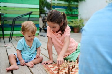 İki çocuk güvertede güneş ışığı altında satranç oynuyorlar. Onların sakin açık hava aktiviteleri bağ kurmayı ve erken öğrenme deneyimlerini teşvik eder. Sahne, aile ve gelişim temalarını aktarır.