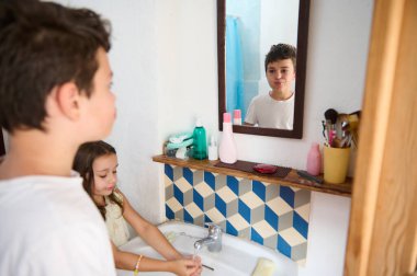 Two children are seen in a bathroom, one washing hands and the other making a funny expression, reflecting daily life and sibling interaction. clipart