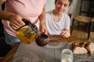 A warm scene showing a parent pouring fresh tea while a child enjoys breakfast. Simple plates and pastries create a cozy atmosphere, highlighting family bonding and morning routines. clipart