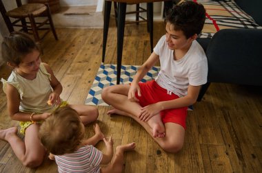 Üç çocuk ahşap bir zeminde oturmuş birbirleriyle etkileşime geçiyor ve eğleniyorlar. Oda atmosferi sıcak, sıcak ışıklandırma ve sade mobilyalarla dolu..