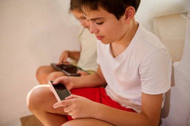 İki çocuk evde oturmuş akıllı telefonlarının keyfini çıkarıyorlar. Görüntü, dijital etkileşimde bulunan, konsantrasyonlarını ve teknolojinin eğlence ve öğrenmedeki rolünü gözler önüne seren çocukları vurguluyor..
