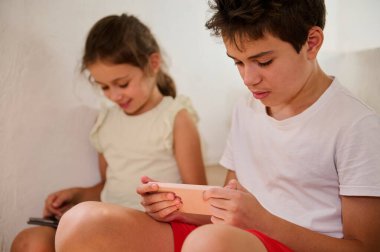 Evde oturan iki çocuk akıllı telefonlarıyla meşgul oluyor, ekranlarına gömülüyorlar, gençlik meraklarını ve teknolojiyi yansıtıyorlar. Dijital etkileşimin sıradan ve sessiz bir anını paylaşıyorlar..